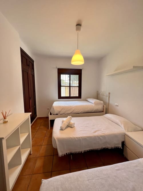 Dormitorio luminoso en Barrosa Home con tres camas, lámpara colgante amarilla, piso de madera y ventana con vista al jardín