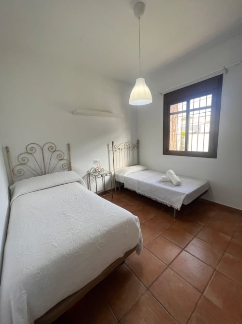 Dormitorio doble con camas blancas, cabeceras de hierro ornamental, luz colgante y suelos de terracota en villa Barrosa Home en España