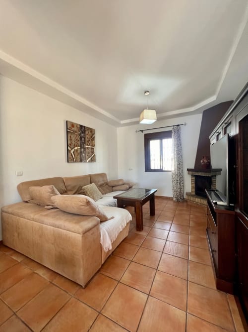 Amplia sala de estar en Villa Pinar del Edén, Barrosa Home con sofá beis, pisos de terracota, chimenea y techo moderno