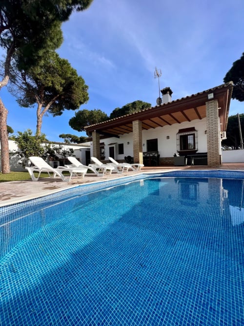 Piscina de aguas cristalinas en villa Barrosa Home con tumbonas blancas, casa de terracota, pinos y cielo despejado