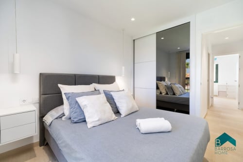 Dormitorio moderno en Barrosa Home con cama tapizada gris, armario espejado, mesita blanca y madera clara