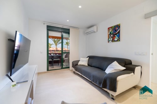 Sala moderna en apartamento Barrosa Home con sofá negro, TV en pared y balcón con vista a palmeras