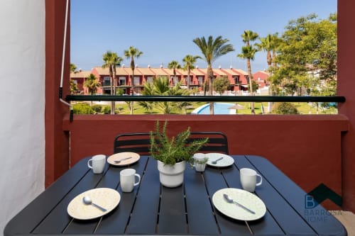 Terraza del apartamento Barrosa Home con mesa de comedor con vistas a palmeras y edificios de tejado rojo en España