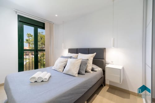 Dormitorio moderno en apartamento Barrosa Home con cama gris, mesillas blancas y puertas de balcón verde con vistas a palmeras