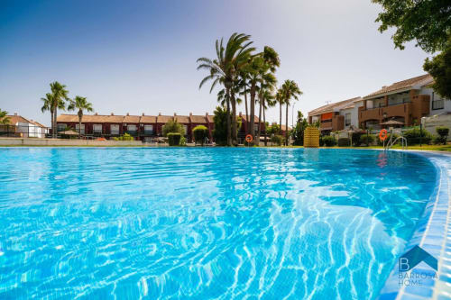 Piscina de aguas cristalinas en el complejo residencial Barrosa Home con palmeras y edificios de terracota bajo cielo despejado
