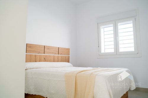 Dormitorio minimalista en Barrosa Home con cabecero de madera, ropa de cama blanca y ventana con persianas con vistas a la costa española