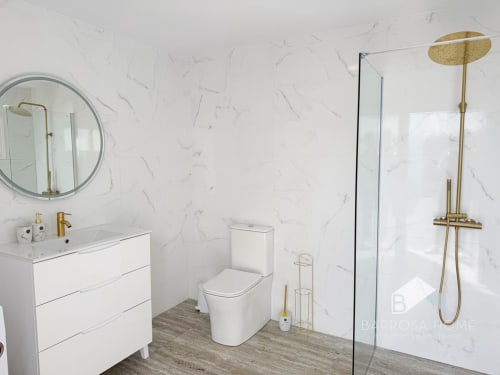 Baño moderno de la villa Barrosa con paredes de mármol, tocador blanco, inodoro, accesorios dorados y ducha abierta