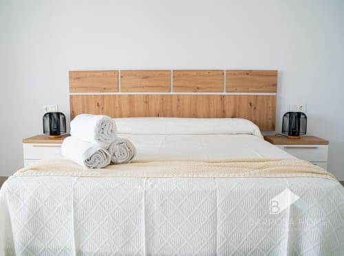 Habitación Villa Horizonte Barrosa Home con cabecero de madera moderno, ropa de cama blanca, toallas enrolladas y lámparas de noche negras