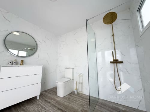 Baño moderno de la villa Barrosa Home con paredes de mármol, griferías doradas, espejo redondo, tocador blanco e inodoro