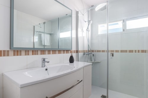 Baño moderno en apartamento Barrosa Home con mueble blanco, espejo grande, ducha de lluvia y cenefa decorativa de azulejos.