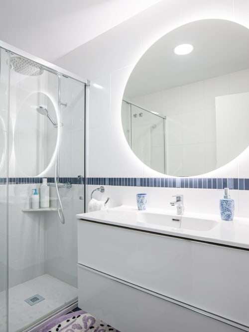 Baño moderno con vanidad blanca, espejo redondo iluminado, ducha de cristal y acentos en azulejos azules