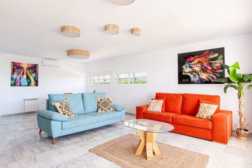 Sala de estar moderna en Barrosa Home con sofá azul, sillón rojo, obras de arte coloridas, luces de techo de madera y mesa de centro de vidrio