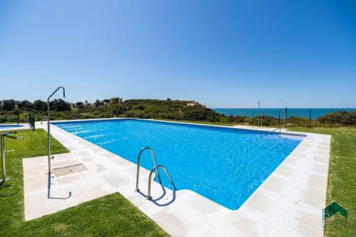 Amplia piscina rectangular con agua azul en el complejo Barrosa Home en España, rodeada de terraza blanca y jardín verde