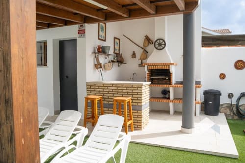 Patio de la villa Barrosa con parrilla de ladrillo, taburetes de madera, tumbonas y decoración rústica bajo pérgola techada