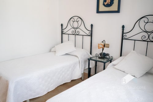 Dormitorio doble en villa Barrosa Home con ropa de cama acolchada blanca, cabeceros metálicos ornamentados y mesitas de noche
