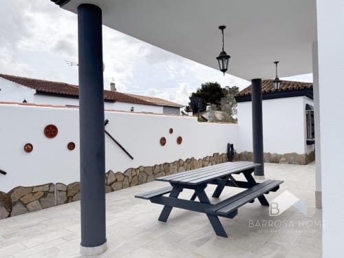 Patio de Barrosa Home con mesa de picnic, columnas oscuras, muro de piedra, lámparas colgantes y arquitectura blanca