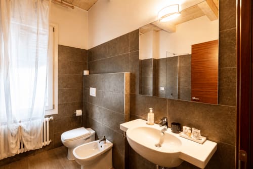 Moderno bagno della Al Battistero Luxury Suite a Ravenna con doppio lavabo, wc, bidet, rivestimenti in piastrelle scure e ampio specchio