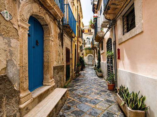 Charmante Kopfsteingasse in Siracusa mit historischen Steingebäuden, blauer Holztür und Topfpflanzen