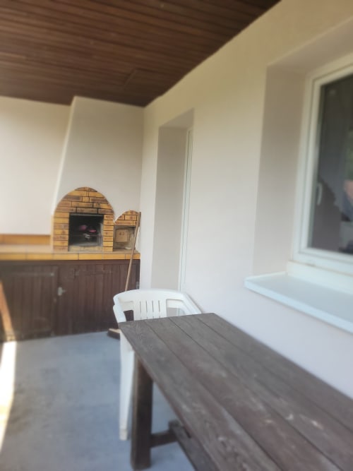 Terrasse couverte avec table en bois, chaise blanche et four à pizza en brique aux Gîtes les Grandes voies au Ménil