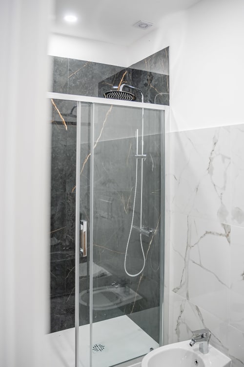 Modern bathroom with glass shower enclosure, dark marble walls with gold veining, rainfall showerhead, and white sink at