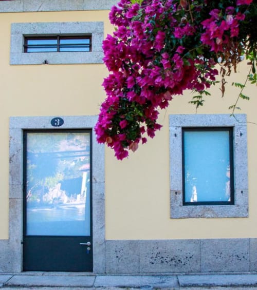 Fachada em tom creme com vibrantes flores de buganvília magenta, janelas com molduras azuis e porta escura na Quinta da