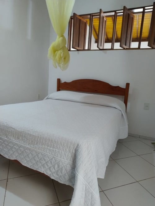 Quarto ensolarado na Casa do Porto com cama de madeira, roupas brancas, cortina amarela e piso de azulejos em Boipeba