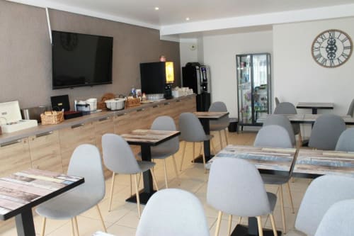 Modern hotel breakfast room at Libre Hôtel in La Vespière with dining tables, grey chairs, TV, and refreshment station