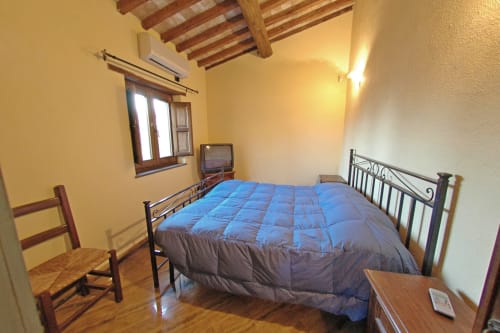 Camera da letto luminosa con piumone blu su letto in ferro, travi in legno, pareti gialle e sedie rustiche all'Agriturismo