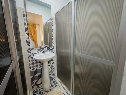 Baño moderno con lavabo de pedestal, azulejos geométricos azules y blancos, espejo y cabina de ducha en el hotel La Puerta del Ángel