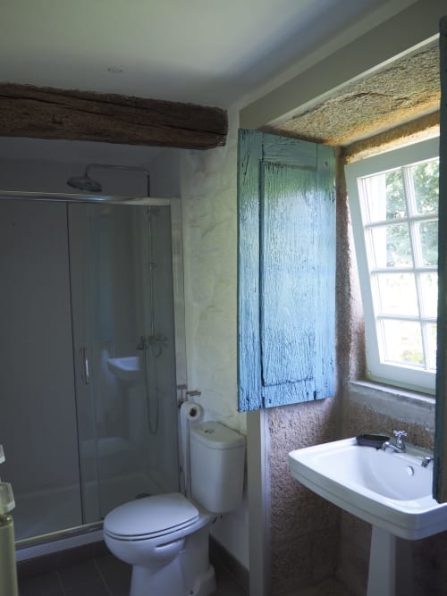 Banheiro moderno com chuveiro, vaso sanitário, pia tipo coluna, armário de madeira pintado em azul, viga rústica e janela na Quinta da