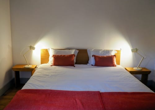 Quarto moderno da Quinta da Coutada com cama branca, almofadas vermelhas, mesas de cabeceira em madeira, luminárias de latão e piso de madeira.