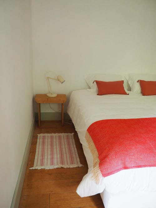 Quarto aconchegante na Quinta da Coutada com cama branca, almofadas vermelhas, criado de madeira e iluminação quente em Arcos de Valdevez