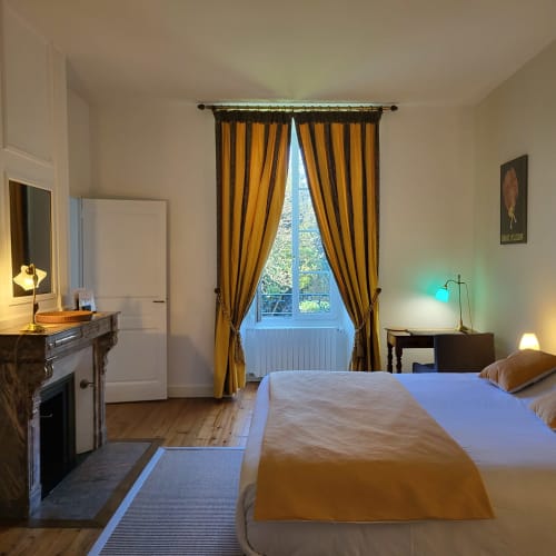 Chambre élégante aux Logis des Tourelles à Cognac avec rideaux dorés, vue sur jardin, cheminée et équipements modernes