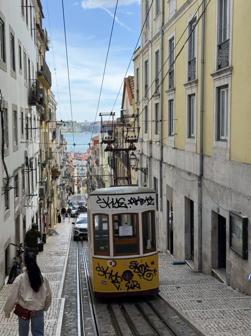 Yellow tram on narrow Lisbon street with historic buildings and river view, near Golden Mermaid Villa