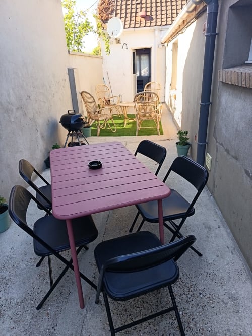 Pink dining table with black chairs in courtyard at Les Meublés d'Hélène, with lounge area and grill visible beyond