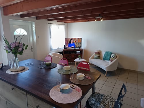 Open-plan living and dining area at Les Meublés d'Hélène apartment, featuring dark wood table, cream sofa, TV, and exposed