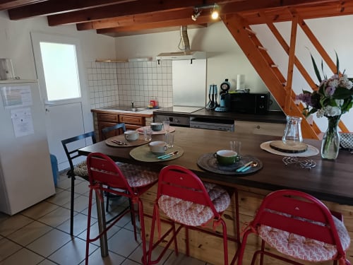 Open-plan kitchen and dining area at Les Meublés d'Hélène with wooden beams, red chairs, and table set for dining.