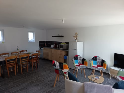 Open-plan living area with wooden dining table, colorful patchwork chairs, kitchen with appliances, and flat-screen TV at Les