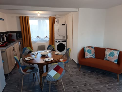 Modern apartment living area with dining table, orange sofa, kitchen, and stacked laundry appliances at Les Meublés d'Hélène