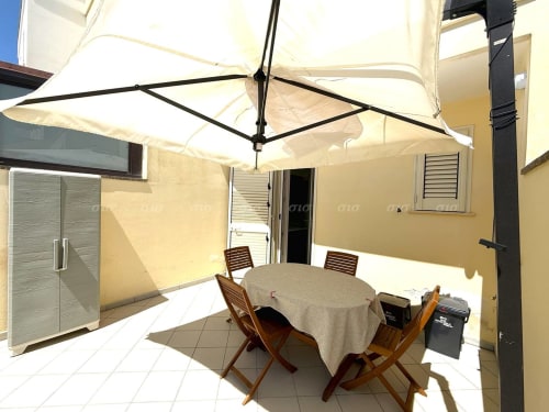 Area pranzo all'aperto ombrellata presso Puglia Estate con tavolo rotondo, sedie in legno e ombrellone color crema a Santa Maria al Bagno