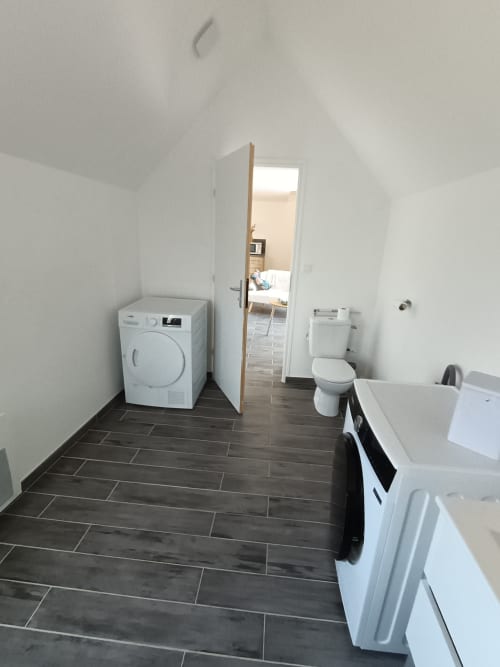Salle de bain moderne avec fixtures blanches, carrelage foncé, lave-linge, toilettes et porte ouverte vers la chambre aux Escardines