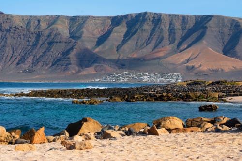 Pristine beach view of Villa Estrella Polar in Tahiche with golden sand, turquoise waters, volcanic rocks, and dramatic cliff