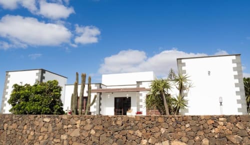 Villa Estrella Polar in Tahiche: Modern white building with stone wall, cacti, and tropical plants under blue sky