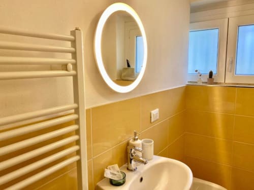 Modern bathroom at The Green Gate Pisa with yellow tile walls, illuminated oval mirror, white sink, and heated towel rack