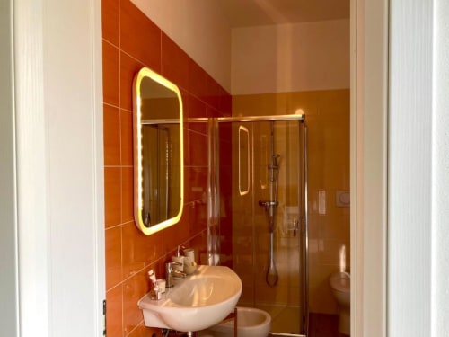 Bagno moderno con piastrelle arancioni, specchio neon illuminato, lavabo a tazza bianca e box doccia in vetro presso The Green