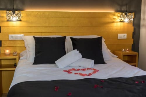 Dormitorio romántico del Hotel Llibrada con almohadas negras, pétalos de rosa roja formando un corazón sobre sábanas blancas, cabecero de madera dorada