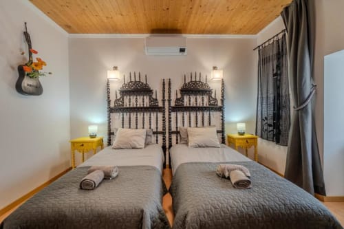 Quarto de hóspedes com duas camas na SOTAM Country House, cabeceiras ornamentadas escuras, criados-de-cabeceira amarelos, teto de madeira e vista para o jardim