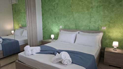 Camera da letto moderna con pareti verde salvia, letto matrimoniale con biancheria bianca e asciugamani arrotolati, comodini con lampade presso Gold Leaf Sicily