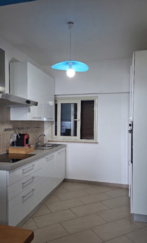 Cucina moderna nell'appartamento Cleopatra con mobili bianchi, lampada a sospensione blu e vista mare da Taormina