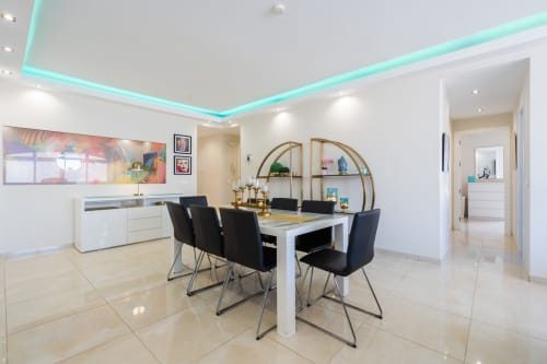 Modern dining and kitchen area in Marbella beachfront apartment with turquoise ceiling lights, white cabinetry, and black